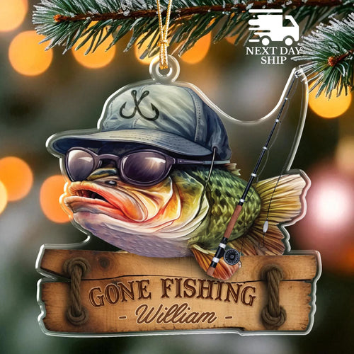 Personalized Christmas ornament featuring a fishing bass wearing a cap and sunglasses, holding a fishing rod, with a wooden sign reading 'GONE FISHING - William -'. Pine branches and warm bokeh lights in the background.