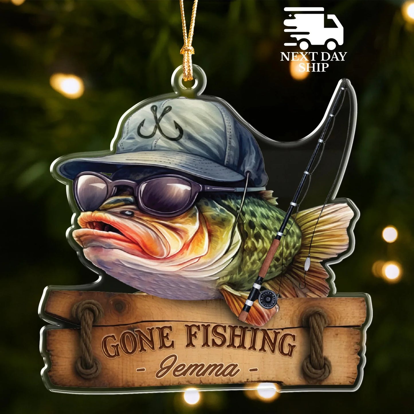 A personalized Christmas ornament shaped like a fishing bass wearing a blue cap and sunglasses, holding a fishing rod, with a wooden sign below reading 'GONE FISHING - Jemma -'. The ornament is shown against a dark, blurred holiday tree background with warm bokeh lights.